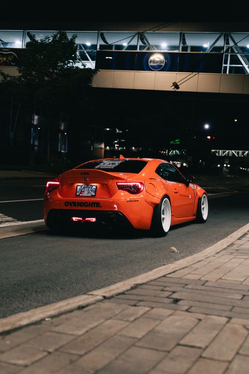 Free Vibrant orange sports car parked on an urban street at night, providing a dynamic cityscape view. Stock Photo