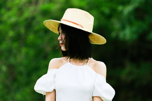 Woman in a stylish straw hat and white dress outdoors amidst lush greenery.