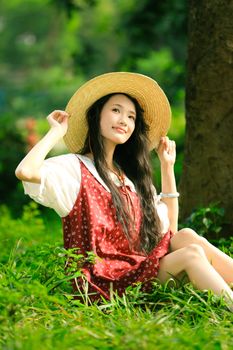 A young woman in a red dress sits on lush greenery, enjoying the sun outdoors.