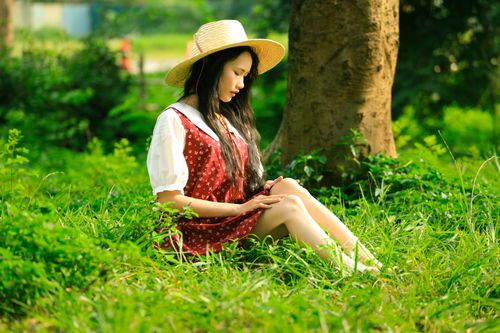 Free A young woman in a red dress and straw hat relaxes on grass under a tree in a Hanoi park. Stock Photo
