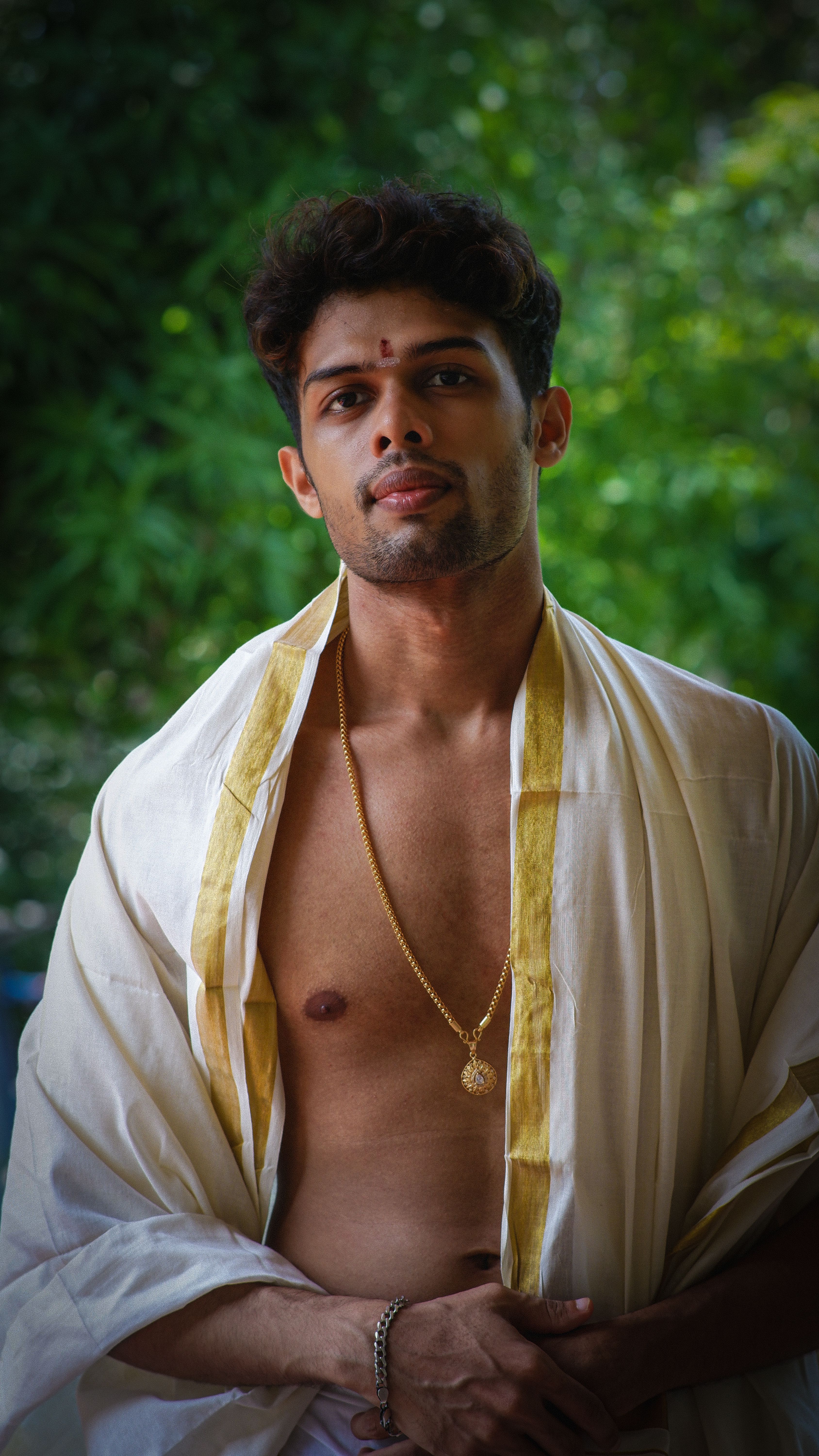 A young man wearing traditional attire stands outdoors, surrounded by lush greenery.