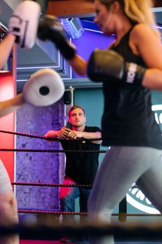 A woman in workout gear trains intensely as a trainer observes in a boxing gym.