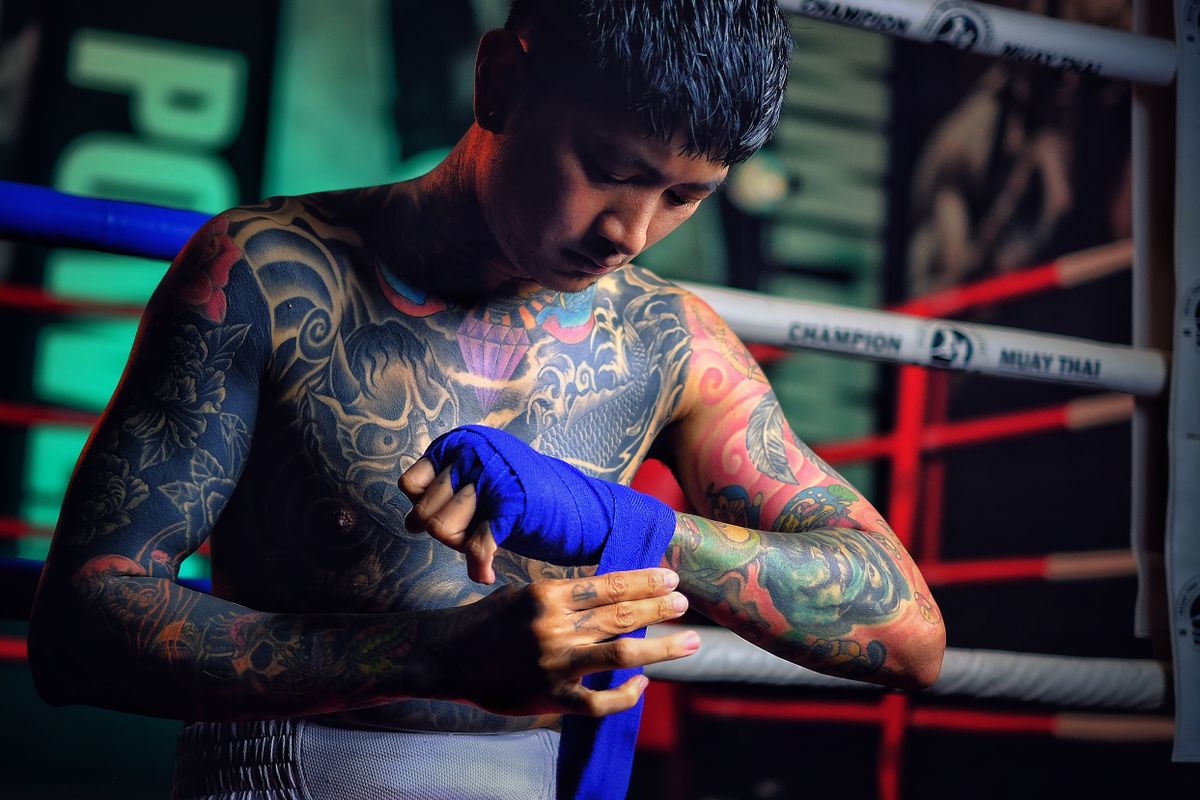 A heavily tattooed fighter wraps his hand with blue bandage against ring ropes reading Champion Muay Thai