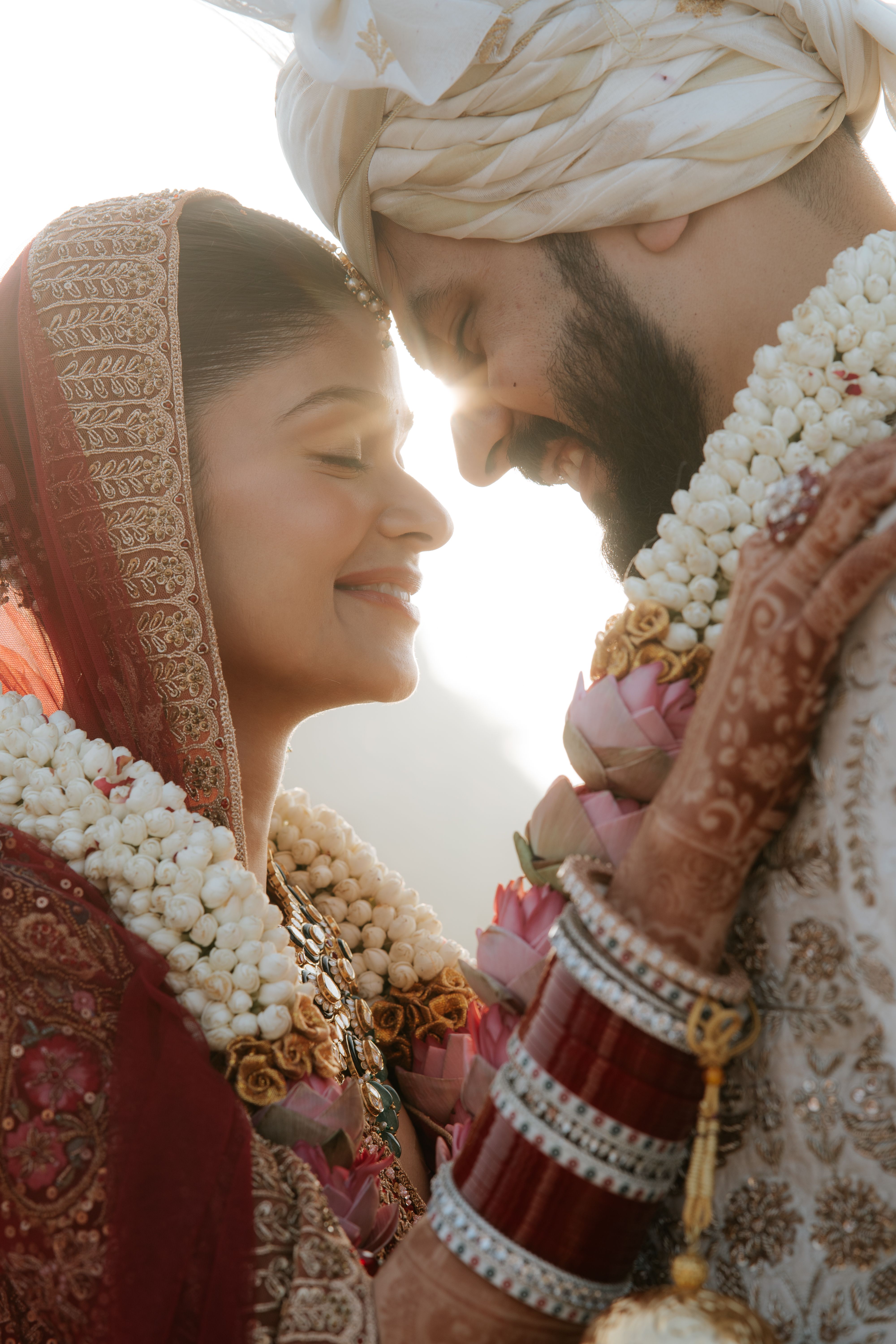 A joyful Indian couple in traditional attire embracing on their wedding day with sunlight in the background.