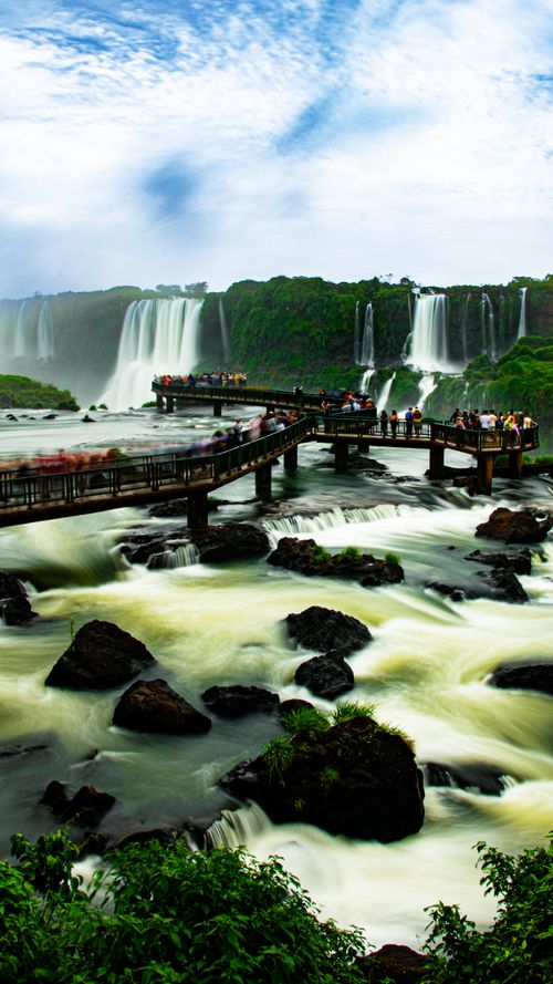 Free Tourists admire the stunning Iguazu Falls from a wooden walkway, surrounded by lush greenery and cascading waterfalls. Stock Photo