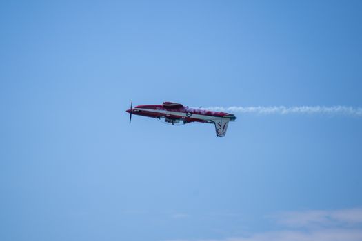 A red aerobatic airplane performs an inverted stunt against a clear blue sky, trailing smoke.