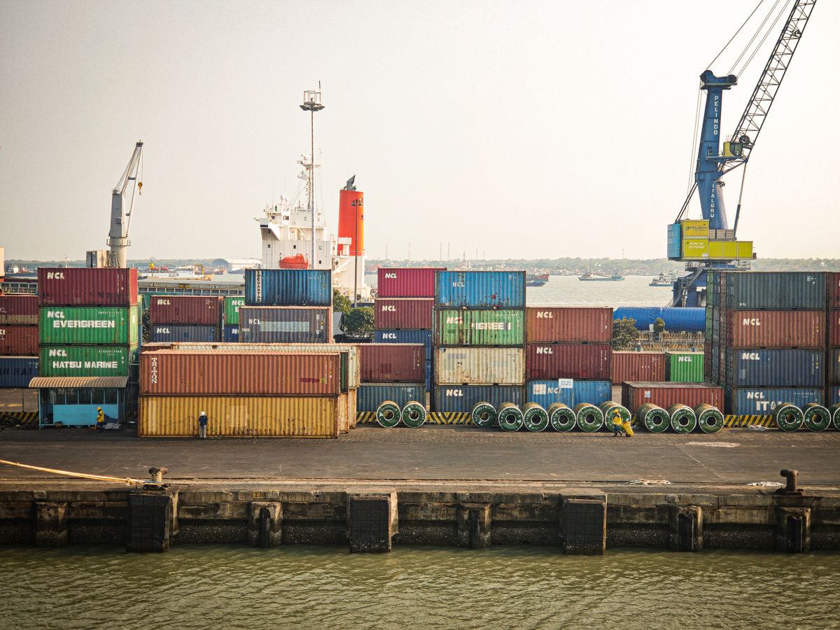 Vibrant shipping containers stacked at an Indonesian port with cranes and a cargo ship in view.