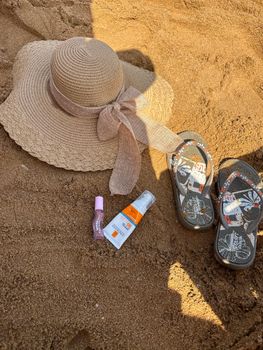 Flat lay of summer beach essentials on sand including hat, flip-flops, and skincare products.