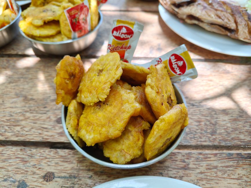 A wooden bowl piled with thin, golden-brown fried plantain chips sprinkled with salt, next to a halved green plantain.