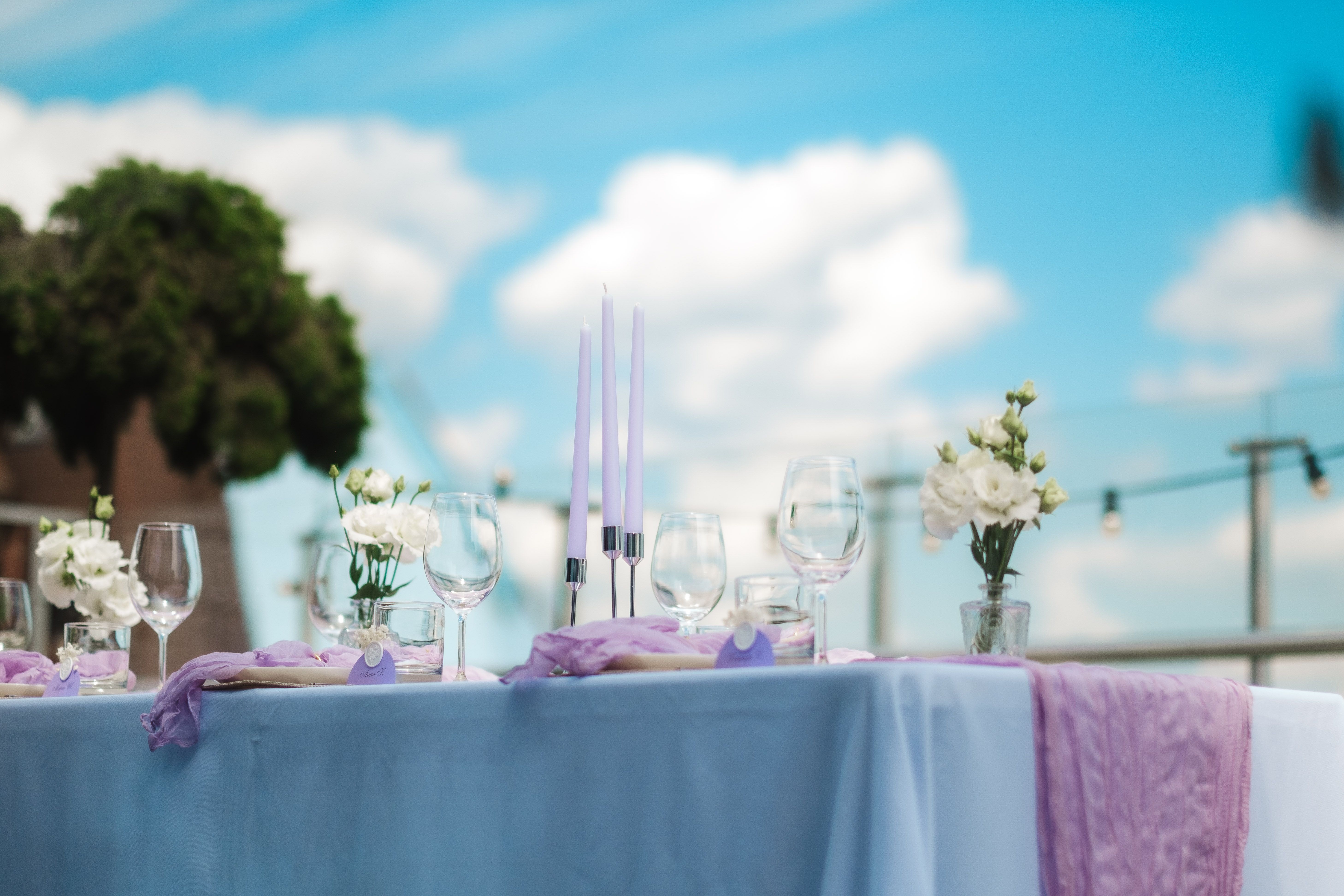 An elegant outdoor table setup featuring lilac accents, floral arrangements, and glassware under a clear blue sky.