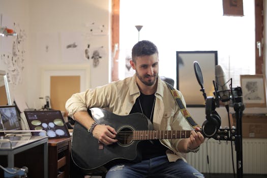 Man playing an acoustic guitar in a home studio with microphones, smiling and relaxed.