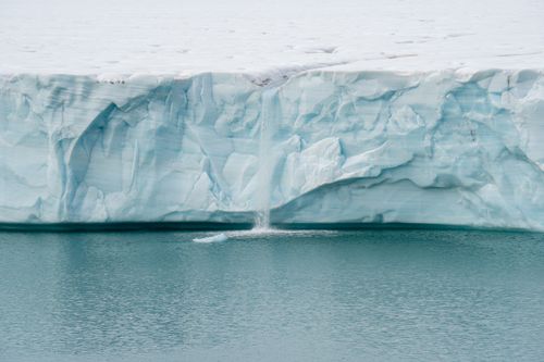 Free A serene view of a glacier edge with meltwater waterfall flowing into a calm ocean. Stock Photo