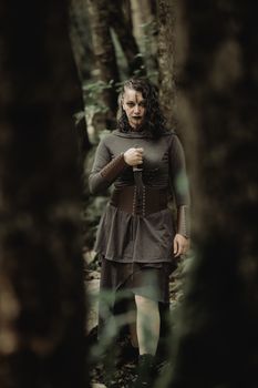 A woman with gothic style stands in a dark, moody forest, holding a knife.