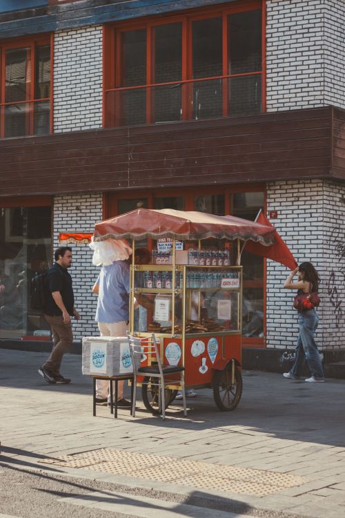 Free A street vendor cart with red umbrella selling snacks on a busy urban sidewalk with pedestrians nearby. Stock Photo