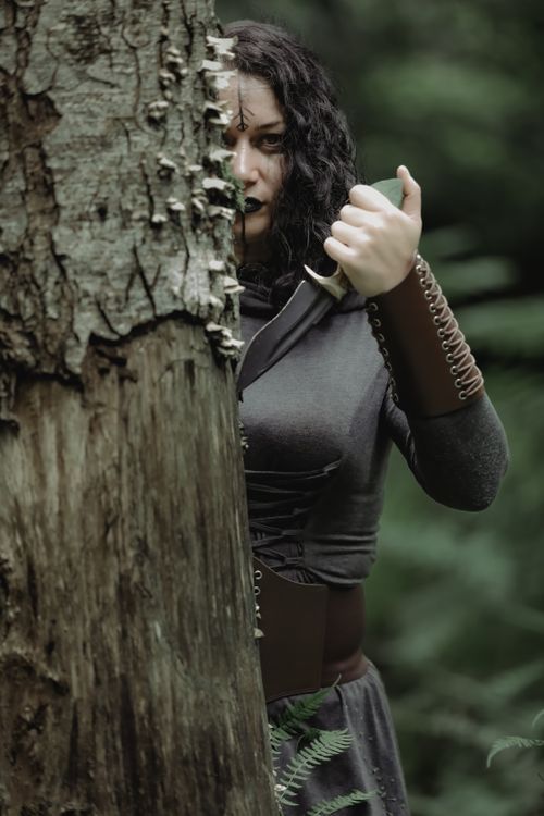 Free A woman in medieval costume holding a dagger behind a tree in a forest. Stock Photo