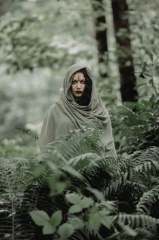 A mysterious figure in a hooded cloak amidst lush green ferns in an ethereal forest.