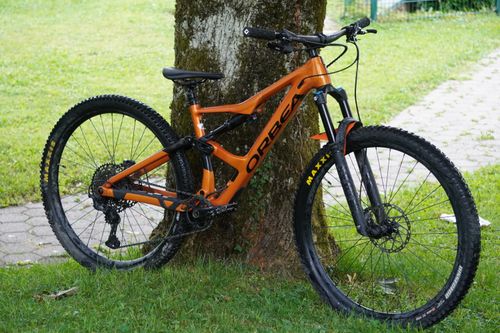 Free A vibrant orange mountain bike rests against a tree, perfect for outdoor adventures. Stock Photo