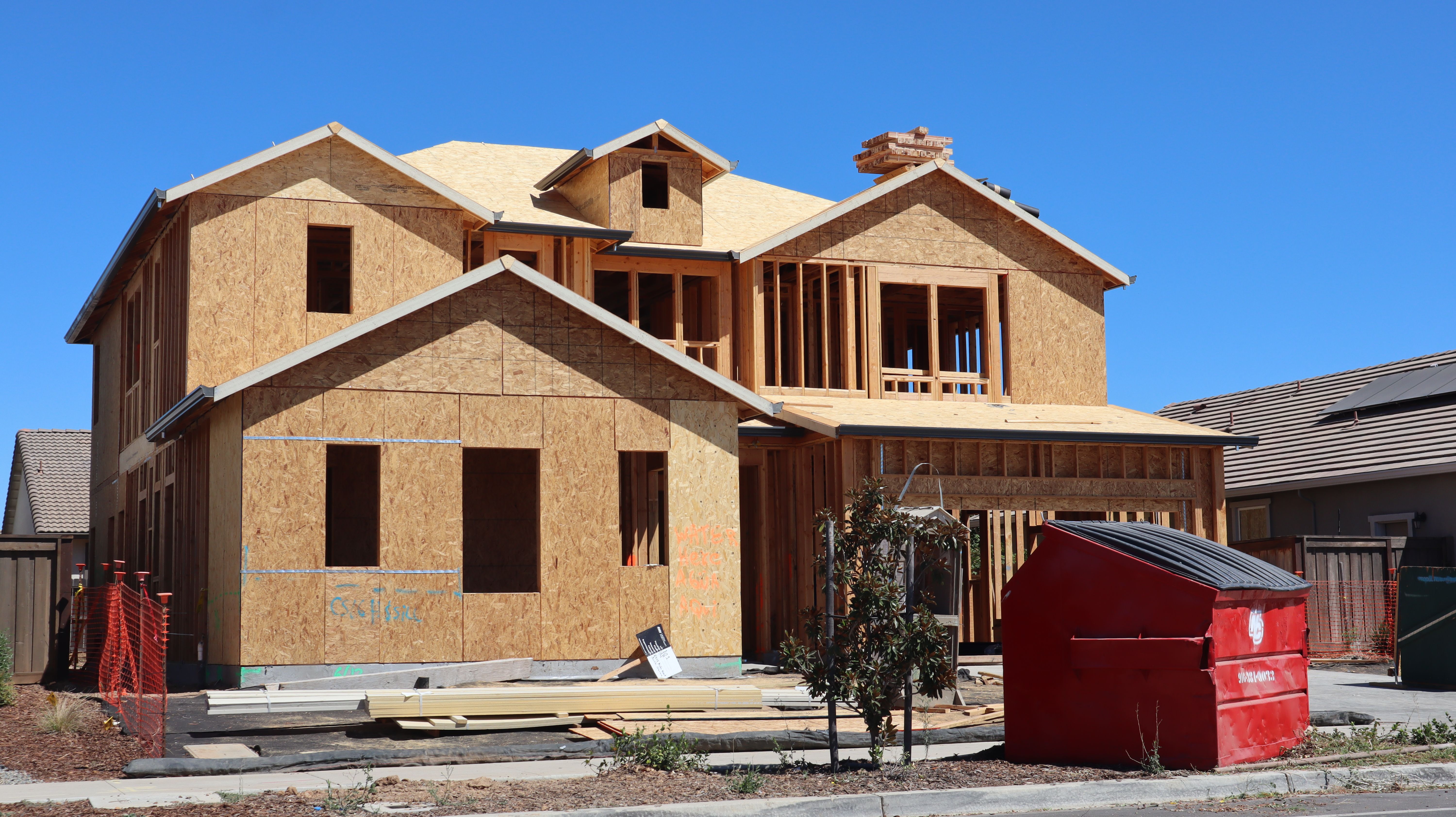 A two-story suburban house under construction, featuring wooden framework and a red dumpster.