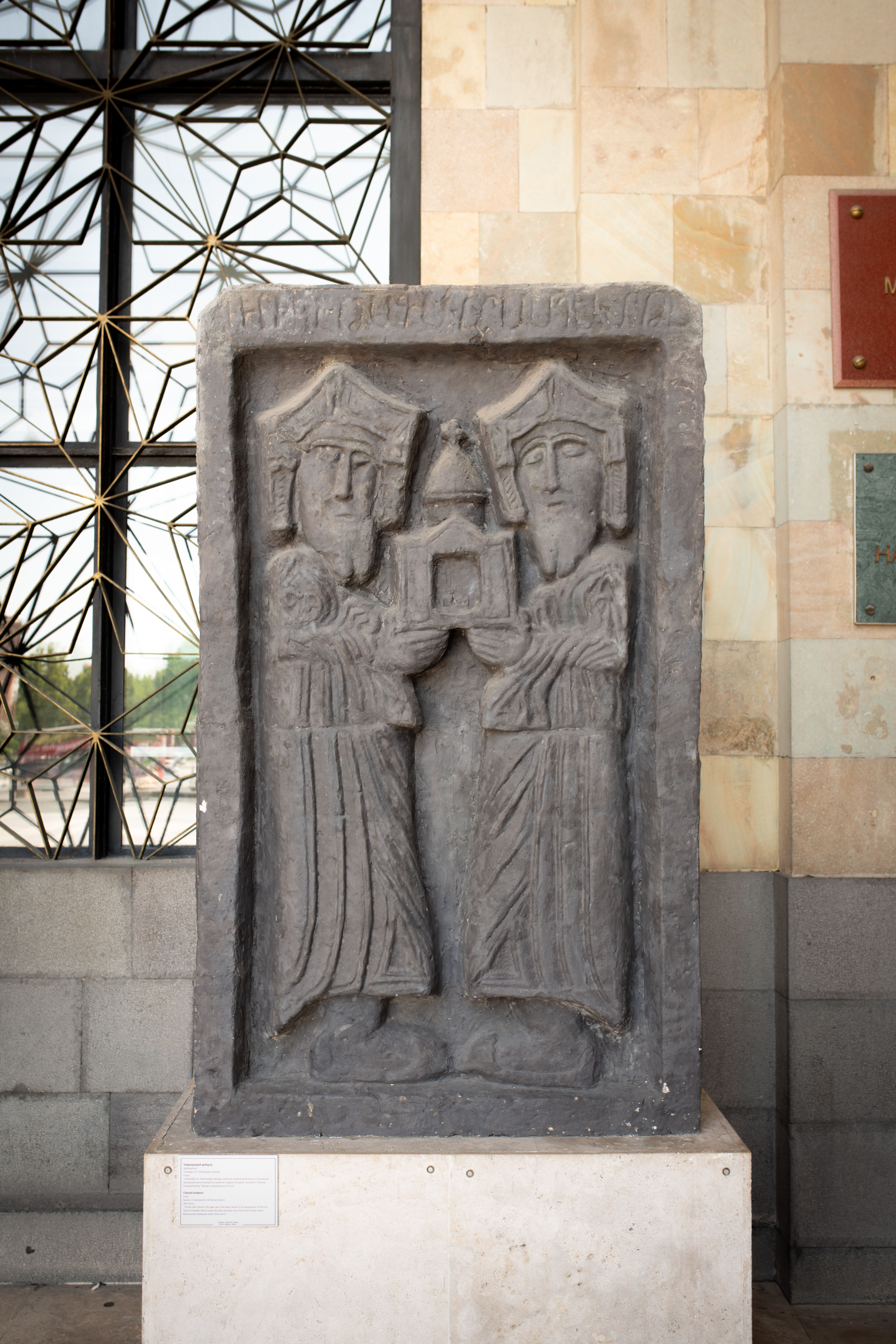 Stone relief artwork featuring two figures holding an object, displayed indoors.