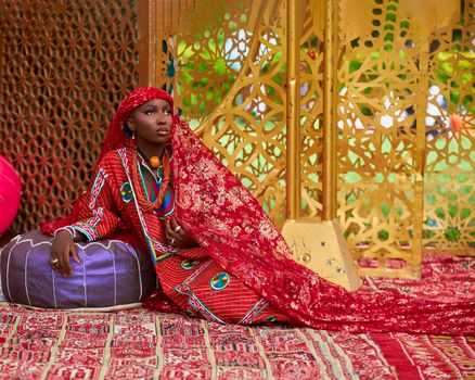 Woman in vibrant traditional Nigerian attire sitting against artistic background outdoors.