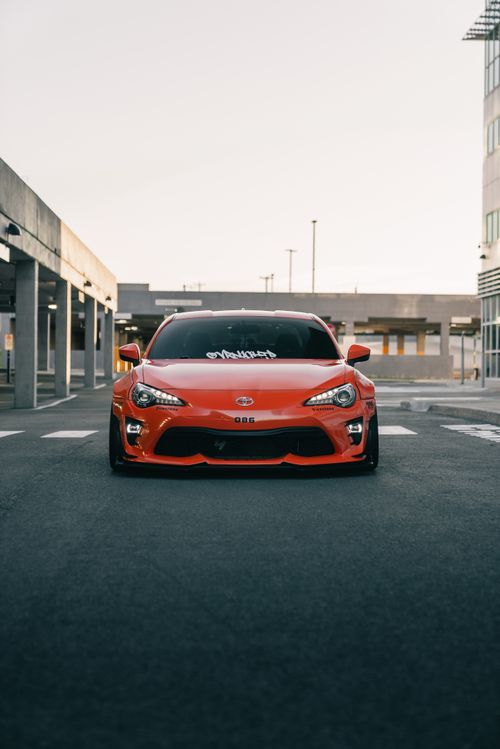 Free A sleek orange sports car parked in a modern urban area under soft daylight. Stock Photo