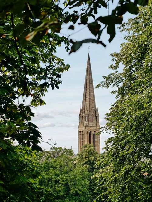 Free Gothic church spire surrounded by vibrant green trees under a clear blue sky. Stock Photo