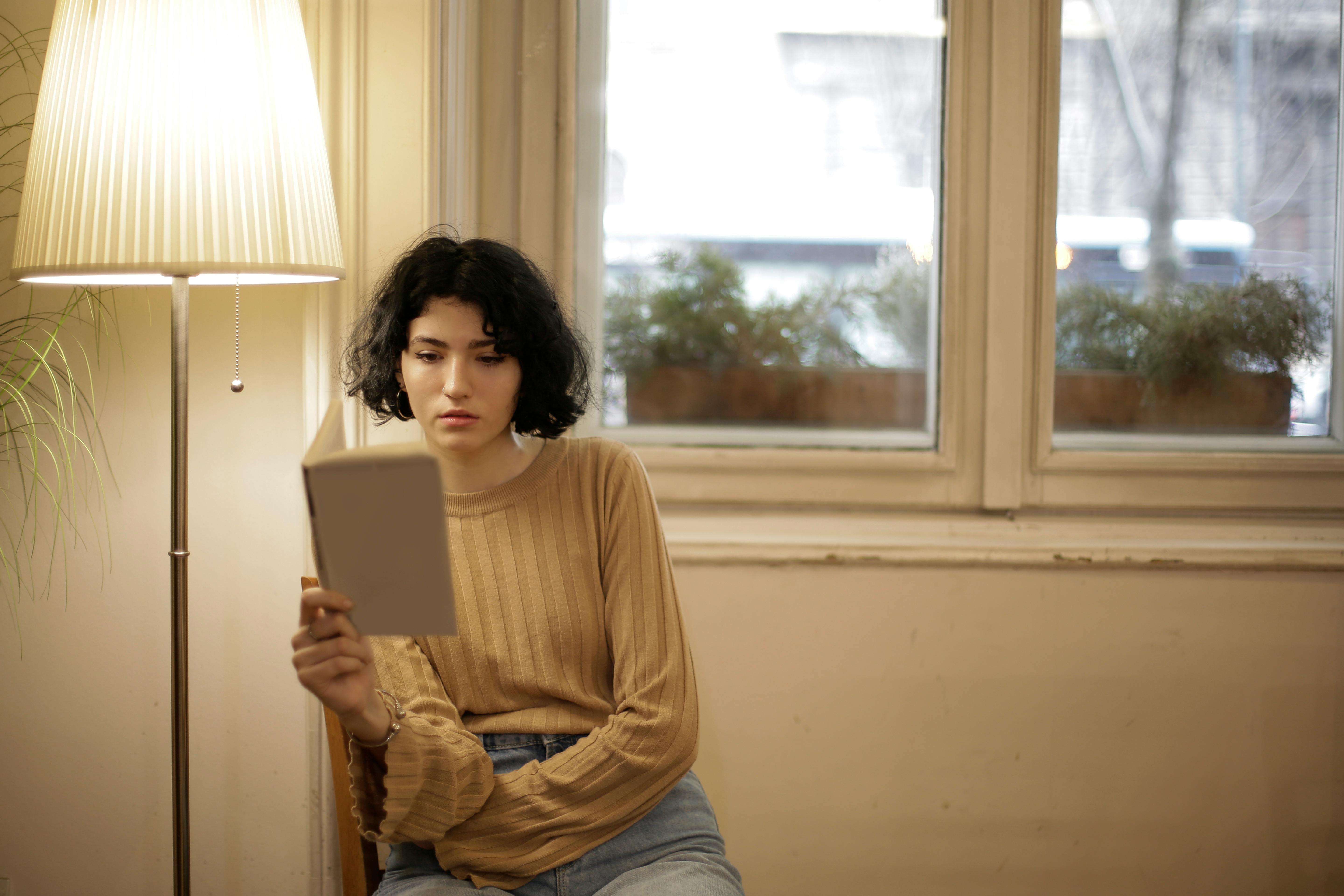 Mulher sentada dentro de casa lendo perto de uma janela sob a luz quente de uma luminária, representando o hábito de leitura para conhecimento sobre o próprio corpo.