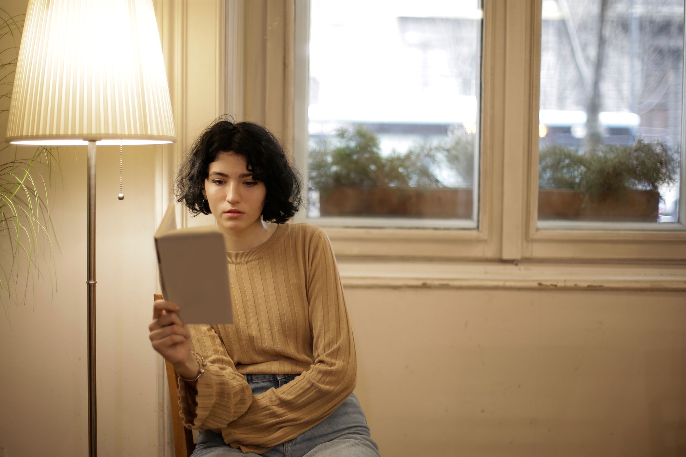Mulher sentada dentro de casa lendo perto de uma janela sob a luz quente de uma luminária, representando o hábito de leitura para conhecimento sobre o próprio corpo.