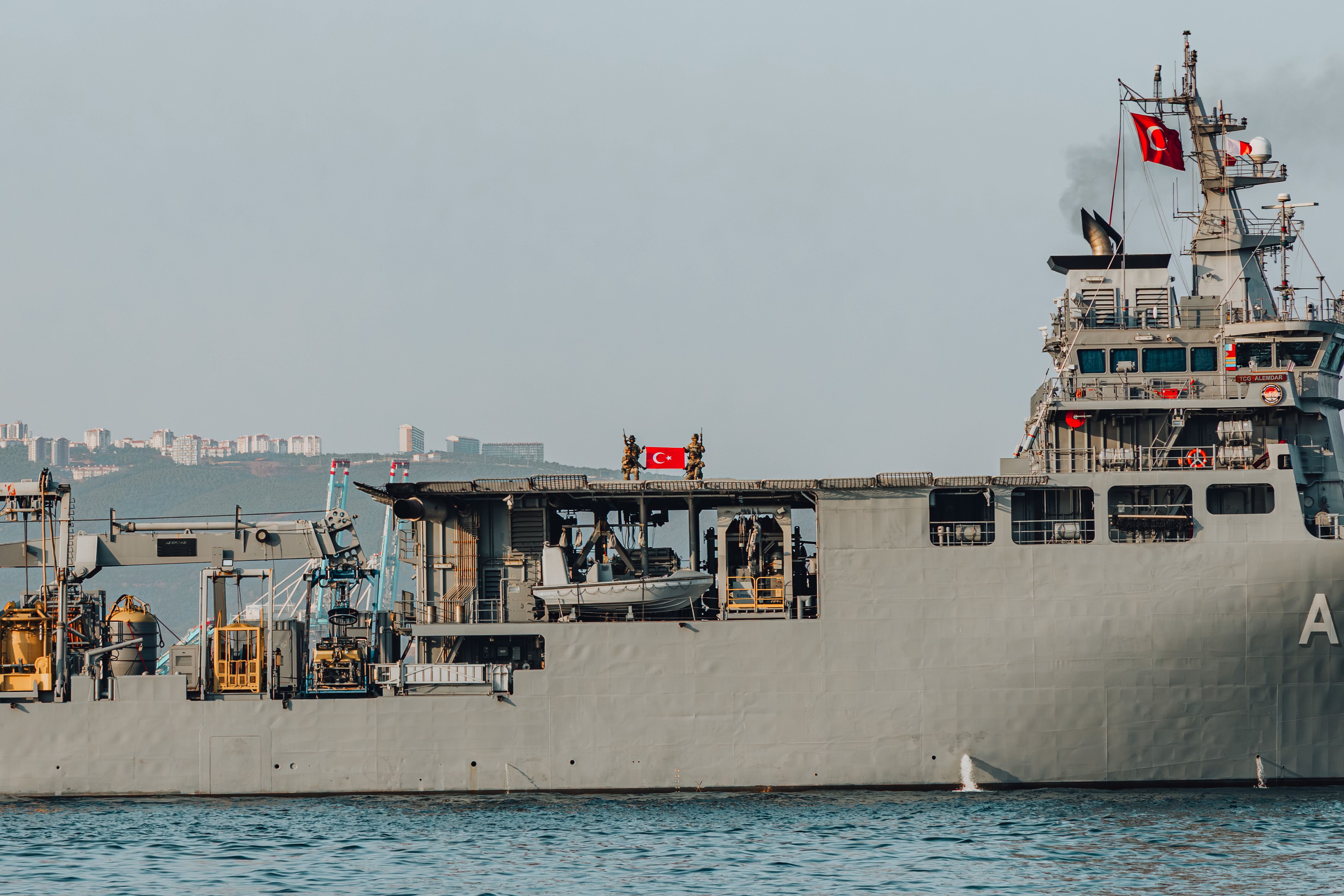 A military naval ship displaying the Turkish flag sails in the sea near an urban skyline.