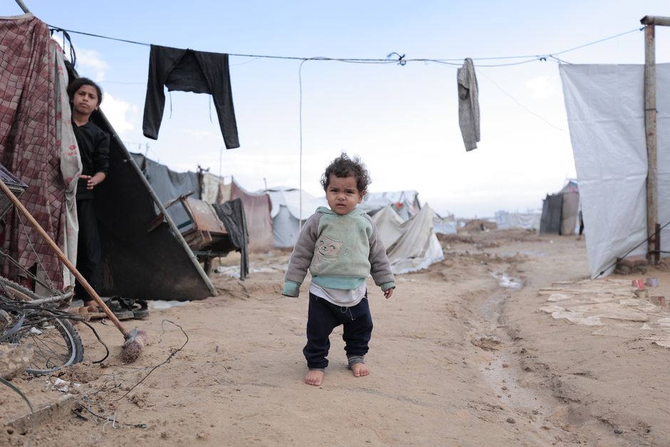 Un niño pequeño camina descalzo en un campo de refugiados, rodeado de tiendas de campaña improvisadas y tendederos de ropa.