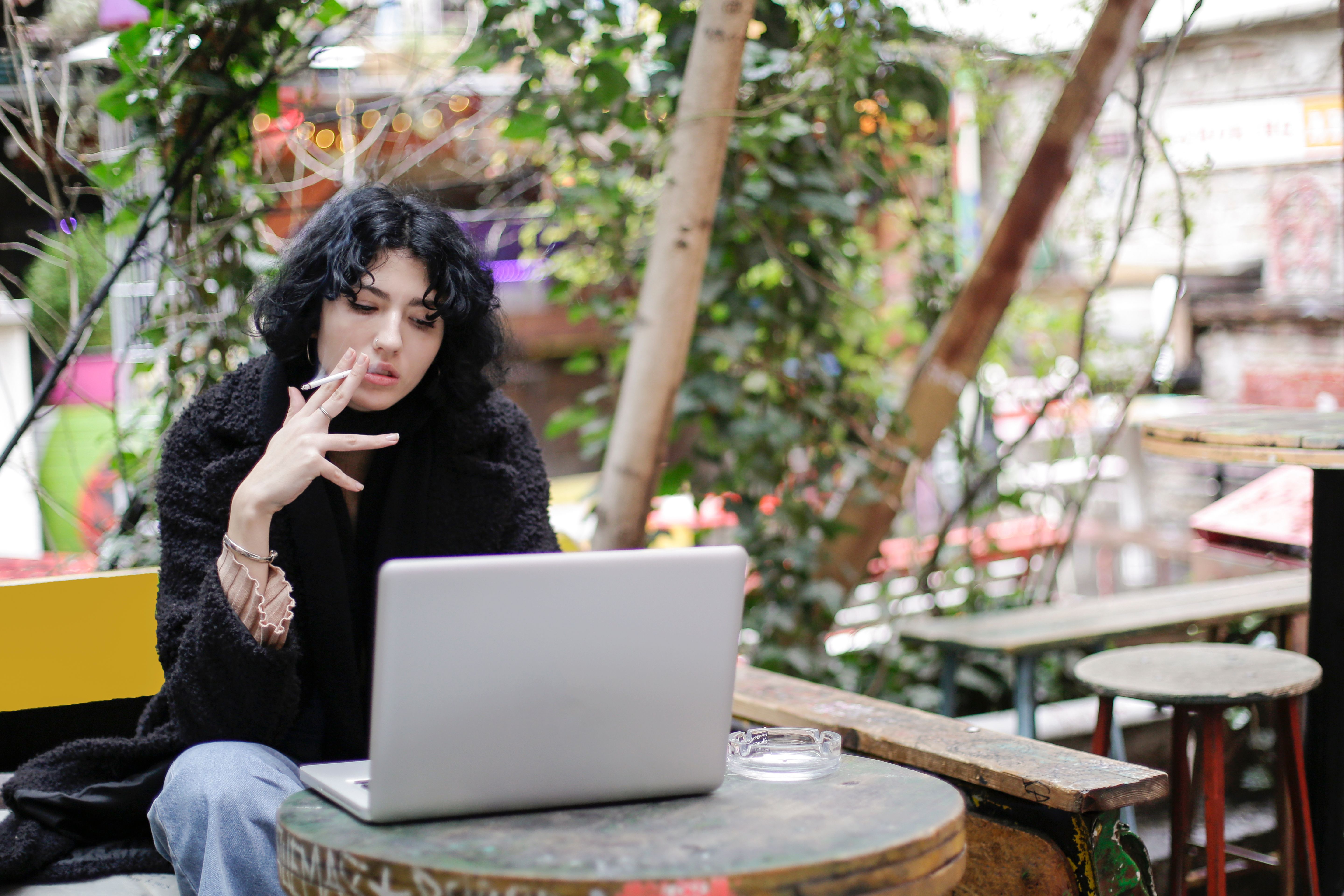 A woman smokes a cigarette while working on a laptop outdoors in a relaxed setting.