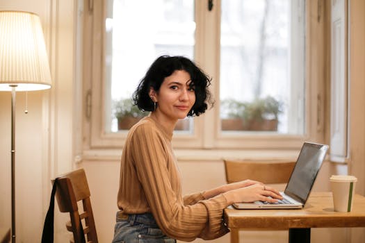 A woman smiling while working on a laptop at home, highlighting remote work and comfort.