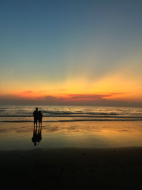 Free A couple stands on Benaulim Beach at sunset, reflecting beautifully on the calm ocean waters. Stock Photo