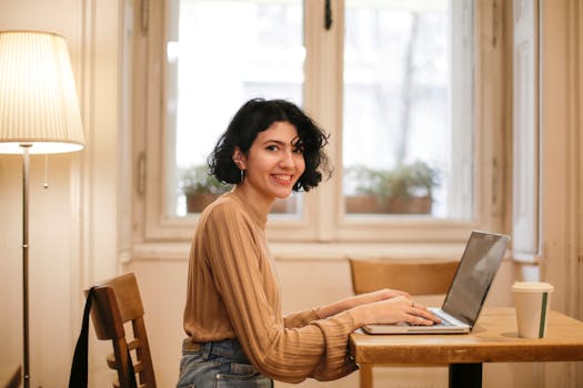 A woman with short hair works on a laptop at a wooden table, smiling. Bright, cozy indoor setting.