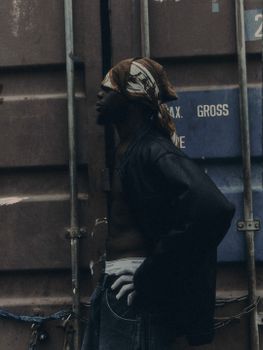 Man in bandana and leather jacket stands by shipping container.