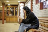 A young woman in a black coat sits on a bench using her smartphone in a well-lit indoor setting.