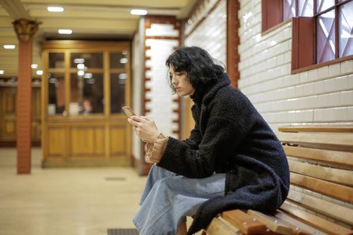 Free A young woman in a black coat sits on a bench using her smartphone in a well-lit indoor setting. Stock Photo