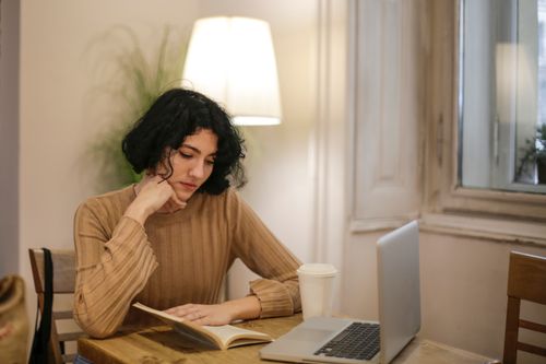Free A woman enjoys reading a book at a cafe table beside a laptop and coffee. Stock Photo