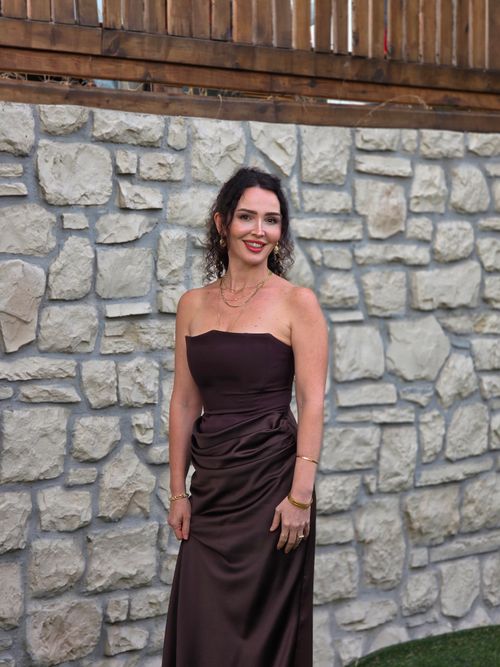 Free Portrait of a woman in a brown dress standing by a stone wall, smiling outdoors. Stock Photo