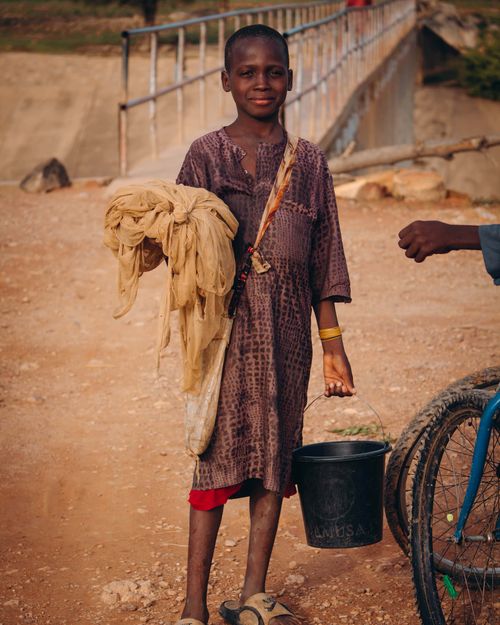Free A young boy stands smiling with a bucket and cloth in rural terrain, near a bridge. Stock Photo