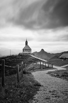 Black and white view of a chapel in Rottach-Egern, surrounded by mountains and dramatic clouds.