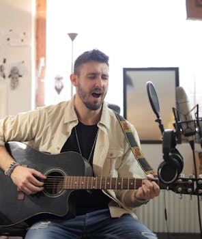 Musician playing guitar and singing into a microphone inside a studio setting.