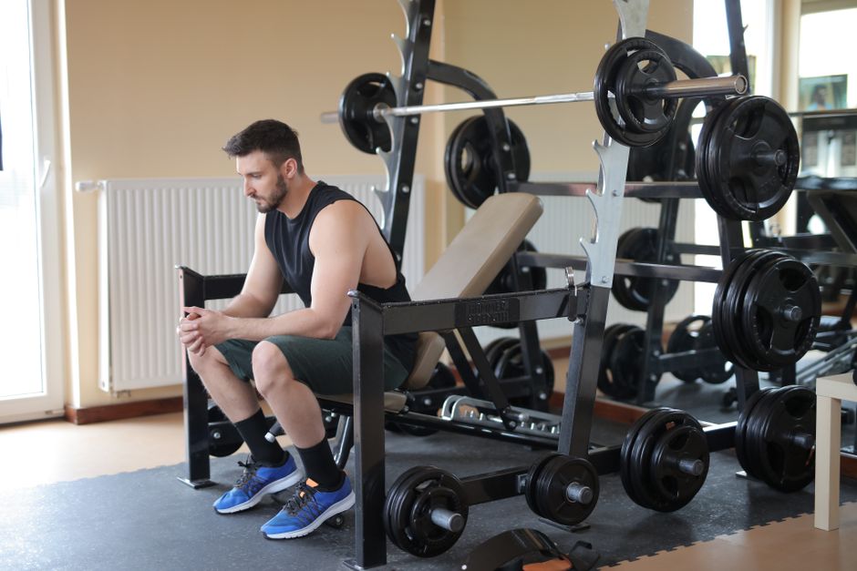 man takes a break at the gym sitting beside a fully loaded barbell, showcasing strength.