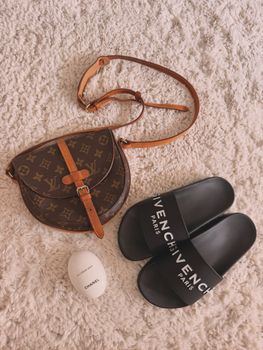 Flat lay of designer handbag, slides, and fragrance on textured surface.