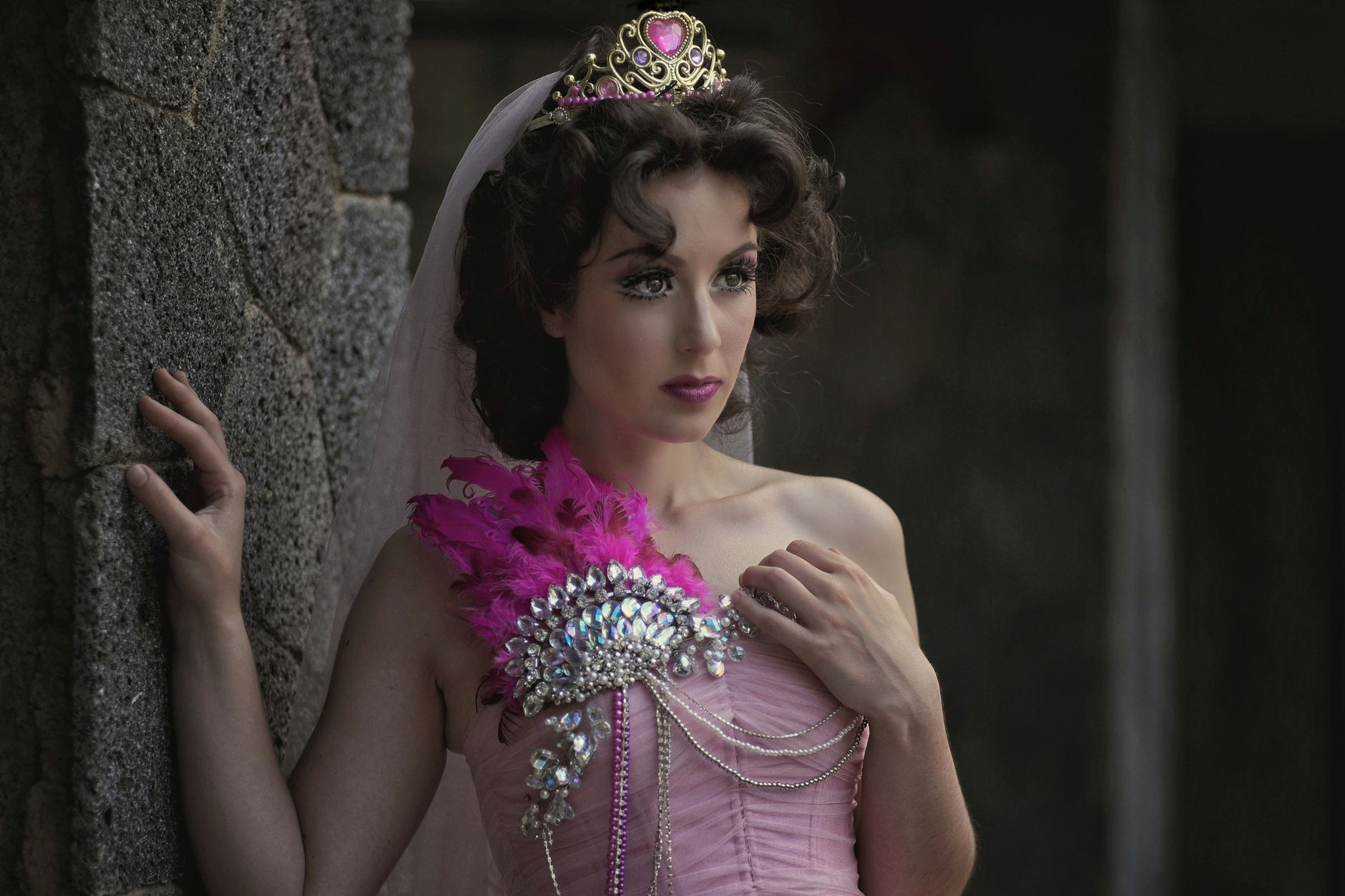 An elegant woman wearing a crown and pink dress adorned with jewels, standing indoors.