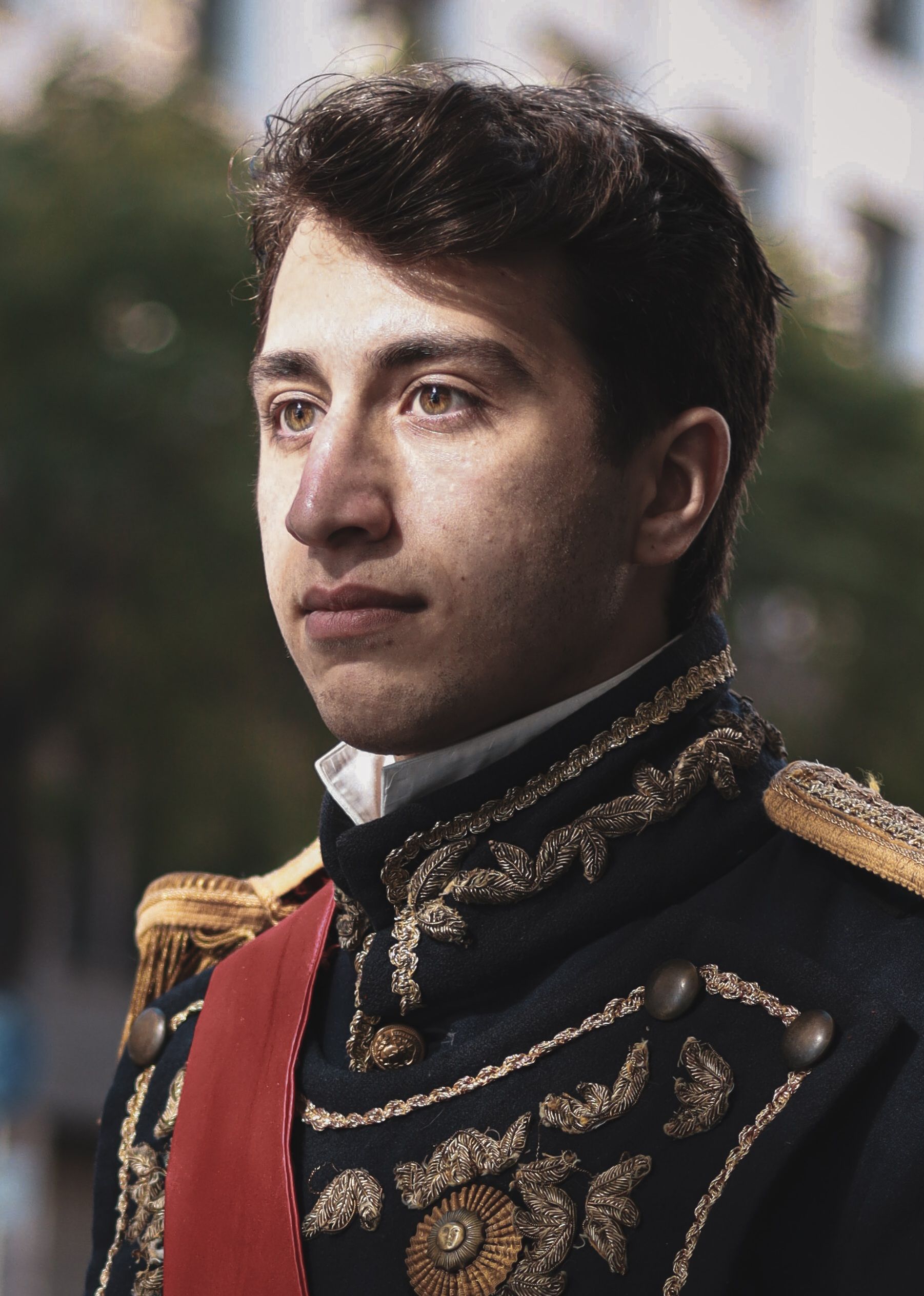 Young man in an ornate historical military uniform looking contemplative outdoors.