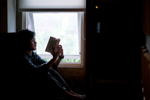 Free A young adult sits by a window, engrossed in a book with natural light creating a peaceful ambiance. Stock Photo