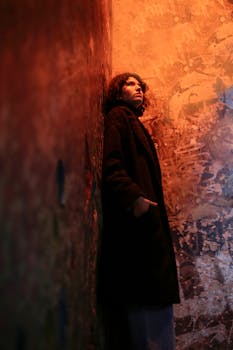 Woman in a black coat leans against a textured wall with dramatic lighting.