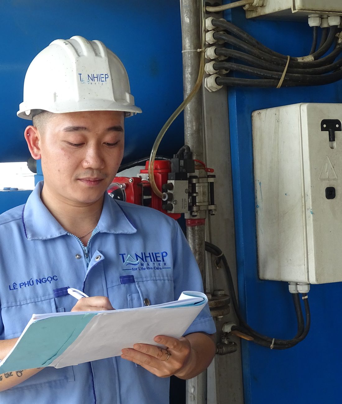 Engineer in a hard hat writing on a clipboard beside a wall-mounted control enclosure.