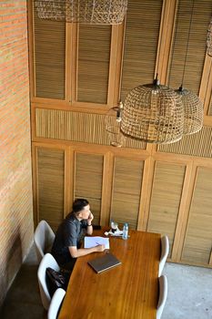 Man sitting at a wooden table in a rustic setting, contemplating ideas with notes and a laptop.